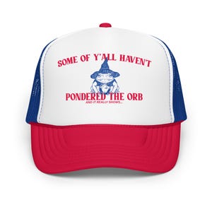 May include: White trucker hat with a red bill and blue mesh back. The front of the hat features a blue and white illustration of a frog wearing a wizard hat and holding an orb. The text "Some of y'all haven't pondered the orb and it really shows..." is printed in red on the hat.