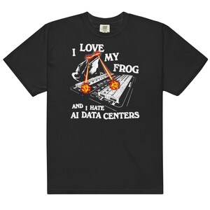 Pode incluir: Uma t-shirt preta de manga curta com um gráfico de um sapo com olhos vermelhos brilhantes a disparar raios laser para um servidor. O texto branco diz I LOVE MY FROG AND I HATE AI DATA CENTERS com explosões de cor laranja.
