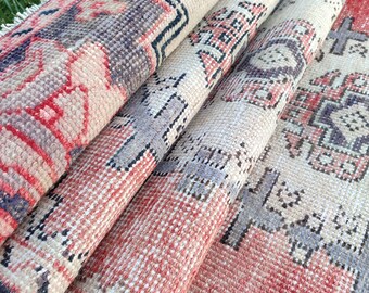 May include: Rolled vintage rugs with intricate geometric patterns in shades of red, beige, and purple. The rugs are displayed on green grass, showcasing their texture and design. The close-up view highlights the detailed weaving and color variations.