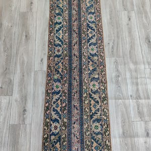 Puede incluir: Una alfombra rectangular estampada con una franja central azul flanqueada por diseños florales y geométricos en tonos azules, beige y rosa. La alfombra tiene un aspecto envejecido y vintage, adecuada para un pasillo o entrada.