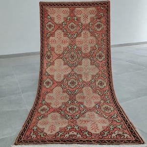 Puede incluir: Una alfombra rectangular con un patrón geométrico en tonos rojos, beige y verdes. La alfombra presenta un diseño central de formas que se cruzan y motivos florales, enmarcado por un borde marrón oscuro. La alfombra se muestra sobre un fondo neutro.
