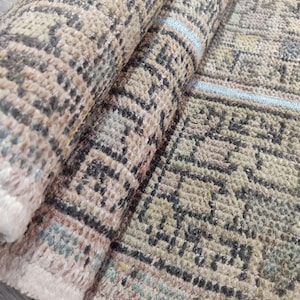 May include: Rolled-up beige and brown patterned rug. The rug features a geometric design with a mix of light and dark brown tones, with accents of blue and pink. The rug's texture appears soft and plush, suitable for home decor.