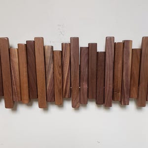 May include: A wooden wall-mounted coat rack with a series of vertical, rectangular wooden pieces of varying shades of brown. The pieces are arranged in a staggered, wave-like pattern, creating a visually interesting design. The rack is mounted on a white wall.