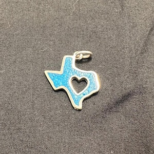 May include: A silver pendant in the shape of the state of Texas, with a heart-shaped cutout. The pendant is filled with turquoise-coloured stones and has a silver border. The pendant is approximately 2.5 cm in length.