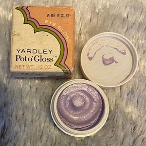 May include: Vintage Yardley Poto'Gloss eye gloss in Vibe Violet. The open container reveals a swirl of lavender-colored eye gloss. The packaging is a small, rectangular box with the product name and net weight of .12 oz.