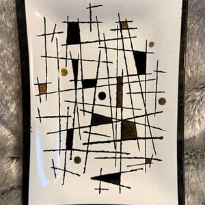 May include: A rectangular glass tray with a white center and a black border. The surface features a black grid pattern of intersecting lines, solid black geometric shapes, and small gold-colored circles and squares scattered throughout the design.