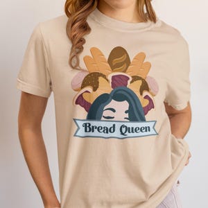 May include: Beige t-shirt featuring a graphic of various breads and a woman's face, with the text "Bread Queen" on a blue banner. The design includes loaves of bread, rolls, and a woman's face with dark blue hair.