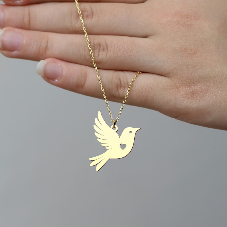 Puede incluir: Un collar de oro con un colgante de paloma. La paloma tiene un coraz&oacute;n cortado en su pecho.