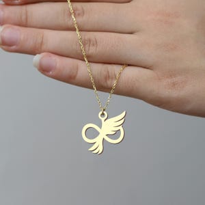 May include: A gold-colored necklace with a pendant shaped like an infinity symbol with wings. The chain is delicate and the pendant is smooth. The necklace is displayed against a neutral background.