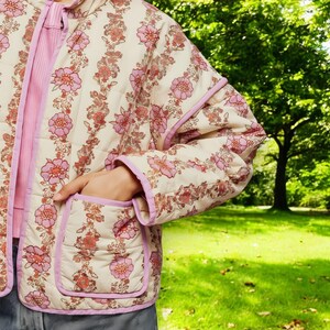 May include: A cream-coloured quilted jacket with a pink floral print and a pink zip. The jacket has two pockets on the front and a pink trim around the edges.
