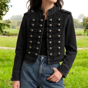 May include: Black denim jacket with a mandarin collar and multiple rows of large, round, silver-tone buttons. The jacket has long sleeves and is paired with blue jeans. The background is a green field and trees.