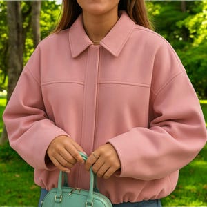 Peut inclure: Veste rose clair avec un col et deux poches avant. La veste est portée avec un jean bleu et un petit sac à main vert clair. L'arrière-plan est un parc vert avec des arbres.