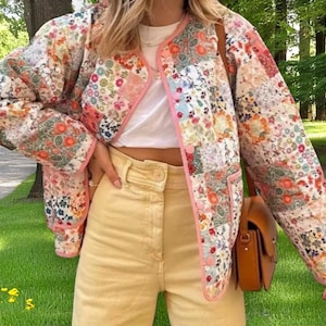 May include: A pink and white floral quilted jacket with a light brown leather shoulder bag. The jacket has a patchwork design with different floral patterns.