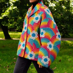 Könnte beinhalten: Eine lebendige, gesteppte Jacke mit Regenbogenmuster und weißen Blumenapplikationen. Die Jacke hat einen klassischen Kragen und einen Knopfverschluss. Das farbenfrohe Design umfasst Rot-, Orange-, Gelb-, Grün- und Blautöne.