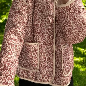 May include: A red and white floral print quilted jacket with a cream coloured trim. The jacket has a zip closure and two patch pockets.