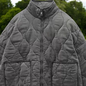 Op de afbeelding: Een grijs gewatteerd jack met een ritssluiting en twee zakken. Het jack heeft een opstaande kraag.