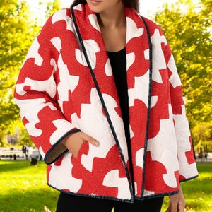 May include: A red and white patterned quilted jacket with black trim. The jacket features a repeating geometric design and has a relaxed fit. The background shows a park with green grass and trees.