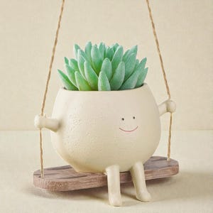 May include: A whimsical off-white ceramic planter shaped like a smiling character, with arms and legs, suspended on a wooden swing. The planter holds a faux succulent with light green leaves. The swing is suspended by natural twine.