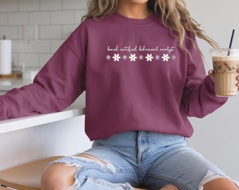 Puede incluir: Una sudadera granate con el texto "board certified behavioral analyst" en blanco, con detalles de margaritas blancas. La sudadera es de manga larga y tiene cuello redondo. La persona lleva vaqueros azules.