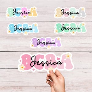 May include: A collection of colorful stickers with the name "Jessica" in various retro fonts. The stickers feature pastel colors like pink, blue, and yellow, decorated with small flowers and hearts. One sticker is held by a hand.