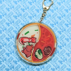 May include: A circular acrylic keychain featuring a split design resembling a yin-yang symbol. One side is cream-colored with a pink ferret and a brown bear, while the other is red with a green vegetable and a brown mushroom. It has a gold metal clasp.