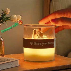 Può includere: Un portacandele in vetro trasparente illuminato con una fascia marrone che recita "I Love you forever". All'interno c'è una foto di una coppia. Le parole "Unique Gift" sono in testo verde. Il portacandele è su un tavolo di legno.