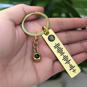 Könnte beinhalten: Goldfarbener Schlüsselanhänger mit einem rechteckigen Anhänger mit einem schwarzen Spotify-Code und -Logo. Der Schlüsselanhänger enthält einen kleinen, runden Anhänger und einen kreisförmigen Schlüsselring. Das Design ist schlicht und elegant.