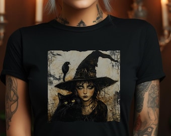 May include: Black t-shirt featuring a witch with a black cat and a raven on her hat. The artwork has a vintage, slightly distressed look. The background is a warm brown with lit candles, creating a mystical atmosphere.