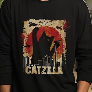 May include: Black long-sleeve shirt featuring a graphic of a black cat silhouette in front of a red sun, with a city skyline and the text "CATZILLA". The design includes Japanese text and airplane illustrations.