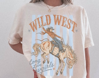 May include: An ivory short-sleeve t-shirt featuring a graphic print of a person riding a bucking horse. The text reads WILD WEST and Long Live Cowgirls. The design includes light blue vertical stripes and orange lettering on the front.