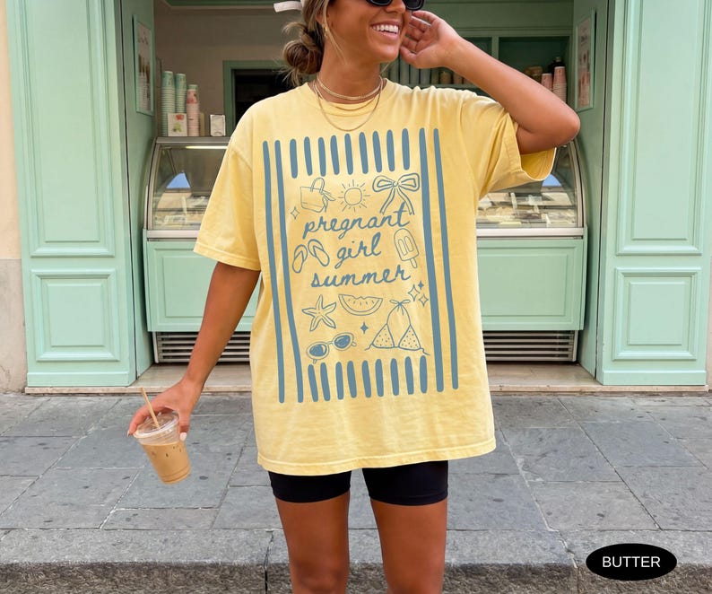 May include: Butter yellow t-shirt with a blue graphic that reads "pregnant girl summer" with summer-themed illustrations. The shirt has a relaxed fit and short sleeves. The person is holding a clear plastic cup with a brown beverage.
