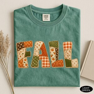 May include: A sage green crewneck t-shirt featuring the word FALL in large, block letters. Each letter is composed of a patchwork design with various autumn-themed patterns, including orange pumpkins, brown leaves, and orange and white gingham checks.