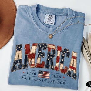 May include: Blue jean colored t-shirt with the word "AMERICA" in a patriotic design. The design incorporates red, white, and blue colors, with stars and stripes. The shirt also features the text "1776 2026 250 YEARS OF FREEDOM" and an American flag.