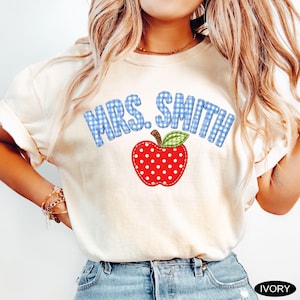 May include: An ivory t-shirt featuring the text MRS. SMITH in a blue and white gingham pattern. Below the text is a red apple graphic with white polka dots and a green and white gingham leaf. The shirt has short sleeves and a crew neckline.