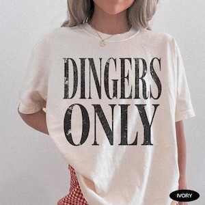 May include: Ivory-colored t-shirt with the text "DINGERS ONLY" in a distressed black font. The shirt has short sleeves and a relaxed fit. The model is wearing a gold necklace and a red and white checkered skirt.