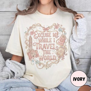 Vintage-inspired pink and beige travel-themed shirt with passport, suitcase, and the quote “Excuse Me While I Travel The World” framed in florals.