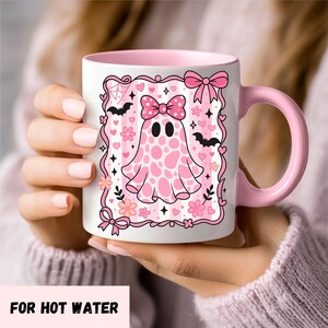 ceramic coffee mug featuring a pink spotted ghost with a bow, surrounded by bats, hearts, and flowers in a coquette aesthetic design