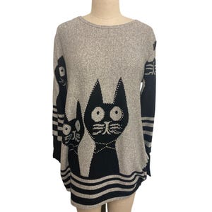 Könnte beinhalten: Grauer Pullover mit einem schwarzen Katzenmuster. Der Pullover hat einen Rundhalsausschnitt und lange Ärmel. Das Katzenmuster umfasst zwei große schwarze Katzen mit weißen Punktdetails und einen gestreiften Saum.