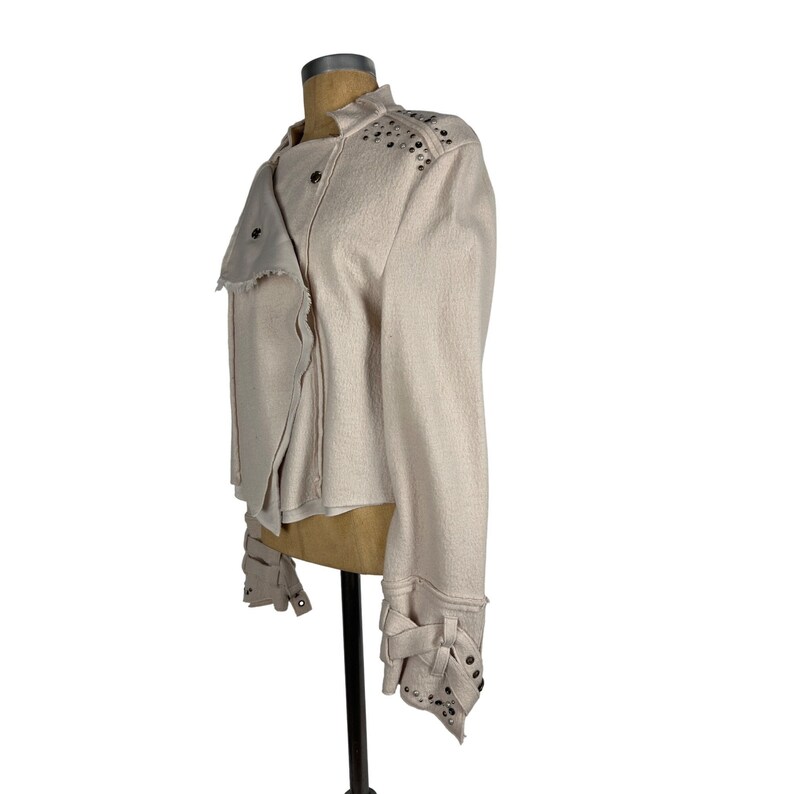 May include: A light beige jacket with a unique asymmetrical design. The jacket features silver-colored studs on the shoulders and cuffs, along with decorative fabric ties. The sleeves have a distinctive cuff detail. The jacket is displayed on a mannequin.