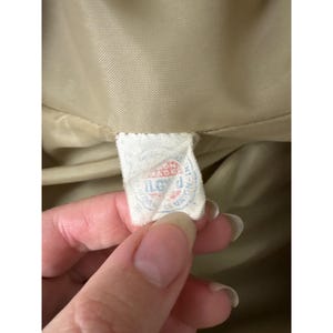 K&ouml;nnte beinhalten: Nahaufnahme eines kleinen, rechteckigen Etiketts mit verblasstem Text und einem rot-blauen Design. Das Etikett wird von einer Hand gehalten, im Hintergrund ist ein beigefarbener Stoff zu sehen. Das Etikett scheint ein Kleidungsst&uuml;cketikett zu sein.