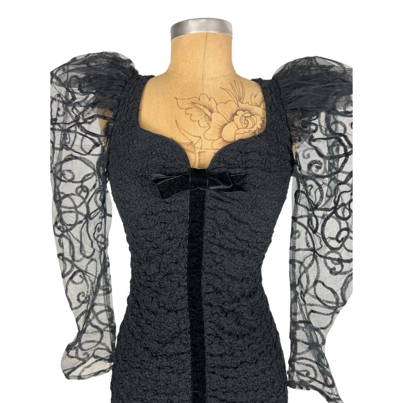 May include: Black dress with a sweetheart neckline and puffed sleeves. The sleeves are made of sheer black lace with a floral pattern. The dress has a velvet ribbon at the neckline.