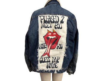 Puede incluir: Chaqueta vaquera azul con cuello de borreguillo color crema. La parte trasera presenta un parche blanco con el texto "PLEASED 2 MEET YOU" y un gráfico de lengua roja con cuernos. Debajo está escrito "GUESS MY NAME".