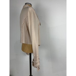 May include: A light beige jacket with long sleeves and a cropped hem. The jacket features decorative stitching and a cuff with buckle closures and silver-colored studs. The jacket is displayed on a mannequin.