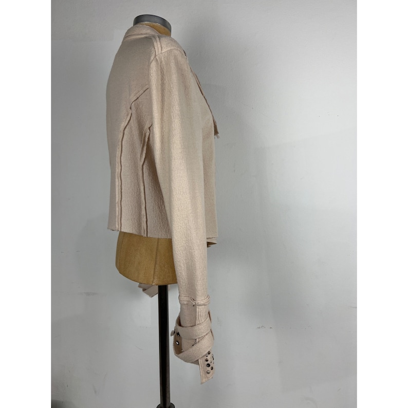 May include: A light beige jacket with long sleeves and a cropped hem. The jacket features decorative stitching and a cuff with buckle closures and silver-colored studs. The jacket is displayed on a mannequin.