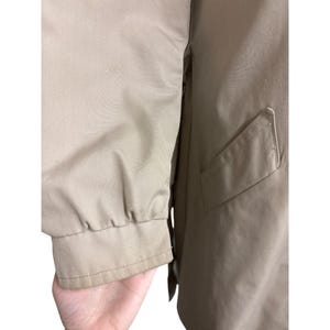 K&ouml;nnte beinhalten: Nahaufnahme eines beigefarbenen Jacken&auml;rmels mit geripptem B&uuml;ndchen. Die Jacke hat eine Tasche auf der Vorderseite und ist aus einem glatten, leichten Stoff gefertigt. Der &Auml;rmel ist leicht zerknittert.