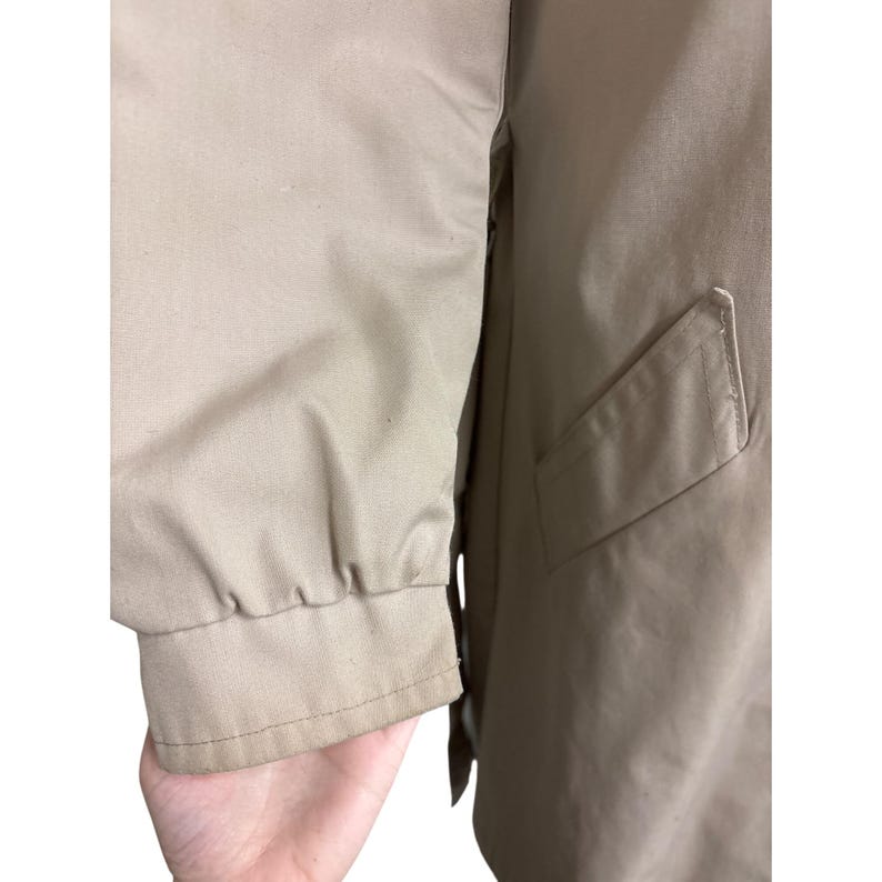 K&ouml;nnte beinhalten: Nahaufnahme eines beigefarbenen Jacken&auml;rmels mit geripptem B&uuml;ndchen. Die Jacke hat eine Tasche auf der Vorderseite und ist aus einem glatten, leichten Stoff gefertigt. Der &Auml;rmel ist leicht zerknittert.