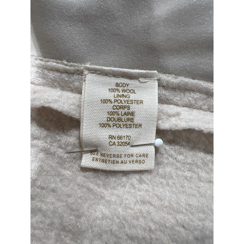 May include: Close-up of a fabric tag with text. The tag reads "BODY 100% WOOL, LINING 100% POLYESTER, CORPS 100% LAINE, DOUBLURE 100% POLYESTER." The tag also includes "RN 66170 CA 32054" and "SEE REVERSE FOR CARE ENTRETIEN AU VERSO."