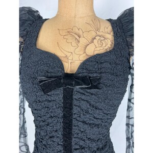 May include: Black lace top with a sweetheart neckline and a velvet ribbon bow. The top has a textured, crinkled fabric and long sleeves.