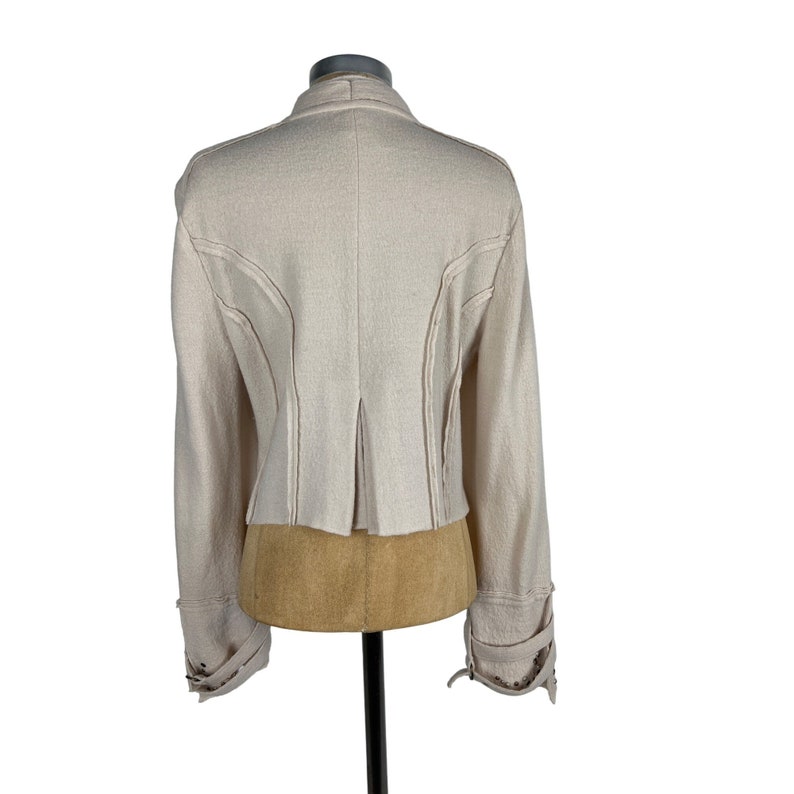 May include: A light beige jacket with long sleeves and a tailored fit. The jacket features decorative stitching along the seams and cuffs, and a pleated back detail. The cuffs have decorative metal accents.