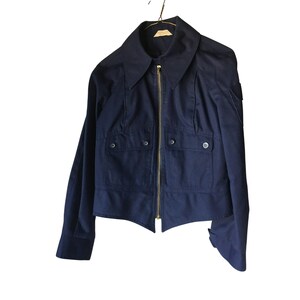 May include: A navy blue jacket with a wide collar and a gold zipper. The jacket has two front pockets with button closures and is hanging on a hanger. The sleeves are long and the jacket appears to be made of a sturdy fabric.
